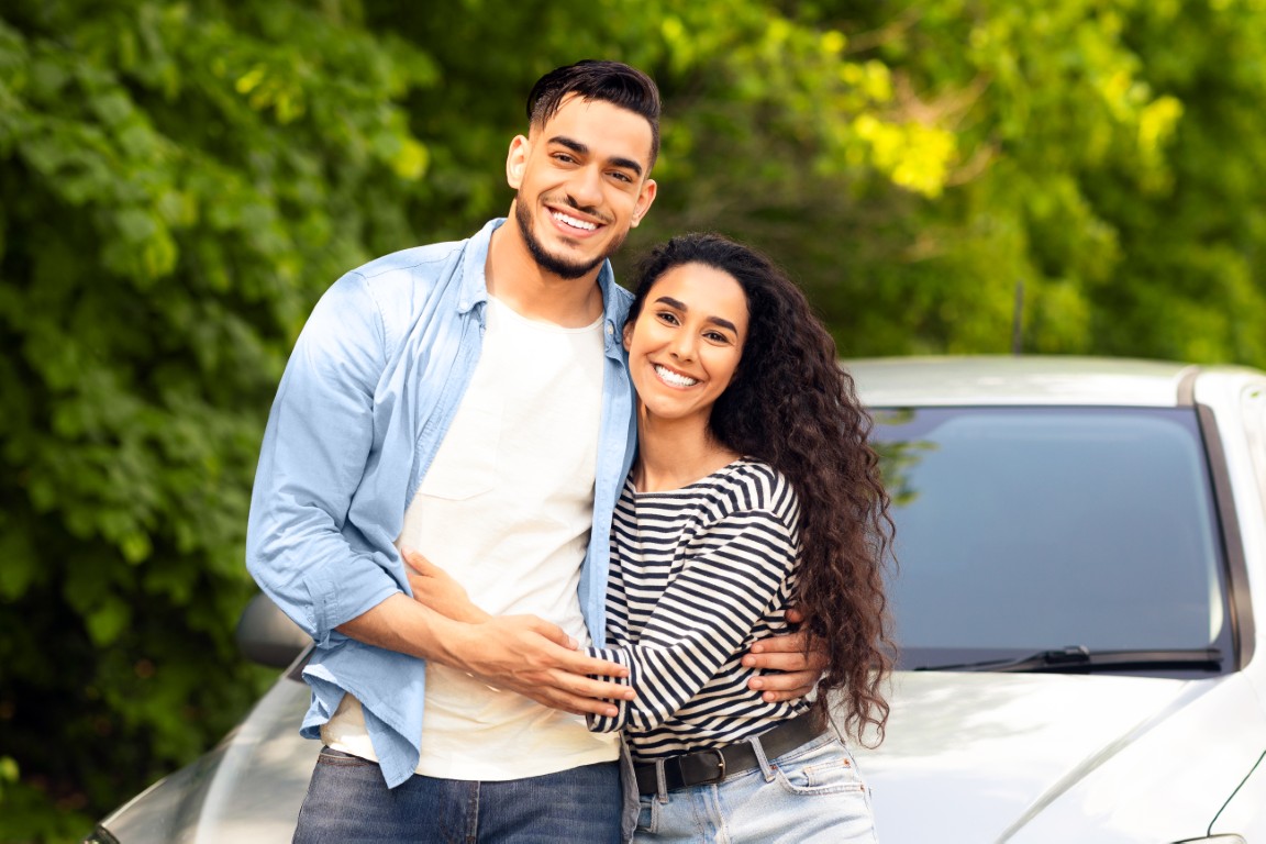 Loving multiracial couple standing by nice auto at countryside, embracing and smiling at camera, copy space. Happy middle-eastern man and woman having car trip together, renting automobile