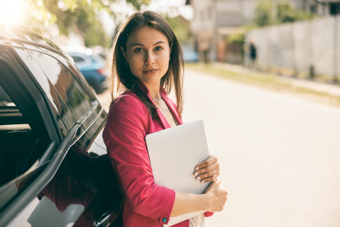 Portrait of young woman holding laptop and leaning to her black car.